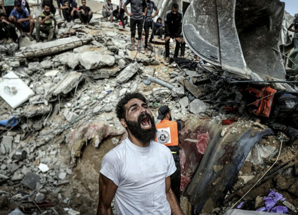 Genocide of Palestinians, man stands in bottom of crater produced by the dropping of a bomb, surrounded by the destroyed remains of peoples houses