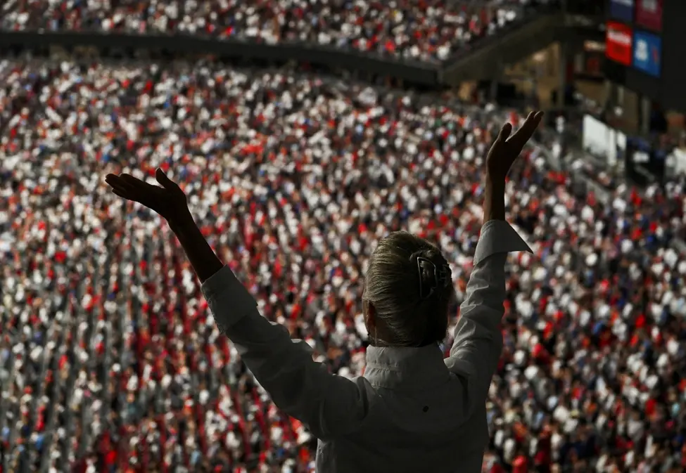 blond white lady raises hands during a prayer at charlie Kirks funeral in front of a 60,000 person crowd. people here supported Kirk's racist and divisive