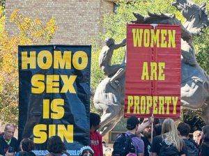 Protests showing two people holding signs that say "Homo Sex IS Sin" and "Women Are Property" a bunch of college age women and men protest around the older people holding signs.