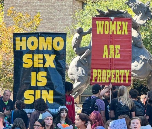 Image showing two men holding signs that say "Homo Sex is Sin" and "Women are Property" while college students protest