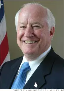 Jim Kolbe smiling at camera in suit and blue tie. with American flag in the background